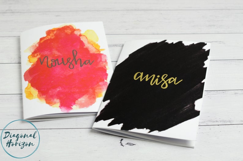 May include: Two small notebooks with watercolour painted covers. The first notebook has a pink and yellow watercolour design with the word "Nousha" written in black. The second notebook has a black watercolour design with the word "amisa" written in gold.