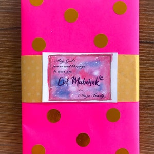 May include: A pink wrapped book with gold polka dots and a yellow gift tag with the text "May God's peace and blessings be upon you Eid Mubarak from Mirza Family".
