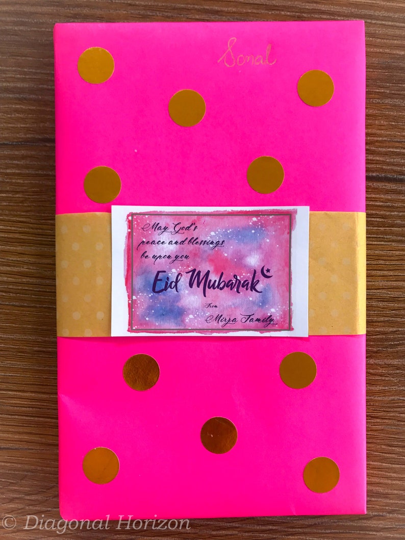 May include: A pink wrapped book with gold polka dots and a yellow gift tag with the text "May God's peace and blessings be upon you Eid Mubarak from Mirza Family".