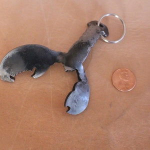 May include: A metal lobster keychain with a silver key ring. The lobster is dark gray with a textured surface and a claw-shaped bottle opener. A penny is next to the keychain for size comparison.
