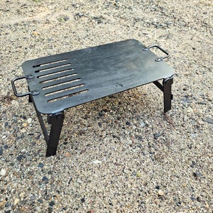 Op de afbeelding: Een zwarte, opvouwbare grill van metaal met een handvat aan elke kant. De grill heeft een rechthoekige vorm en een reeks gleuven die in het oppervlak zijn gesneden.