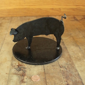 May include: A black metal pig sculpture with a curled tail, standing on a round base. The pig is in a walking pose with its head turned to the side.