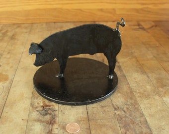 May include: A black metal pig sculpture with a curled tail, standing on a round base. The pig is in a walking pose with its head turned to the side.