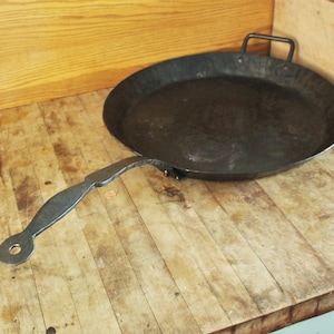 May include: A large, round, black cast iron pan with a long, curved handle. The pan is sitting on a wooden surface.
