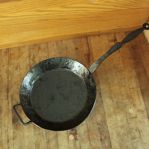 May include: A black cast iron pan with a hammered finish and a long handle. The pan is sitting on a wooden surface.