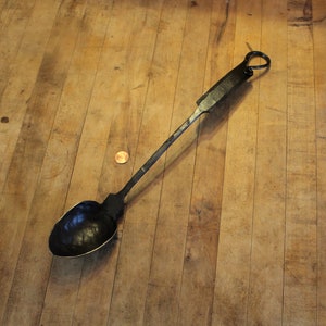 May include: A hand-forged, black metal spoon with a long handle and a rounded bowl. The spoon has a loop at the end of the handle for hanging. The spoon is lying on a wooden surface.
