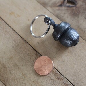 Puede incluir: Un llavero de metal plateado en forma de bellota con un anillo de llave plateado. La bellota es aproximadamente del tamaño de una moneda de 1 céntimo de euro.