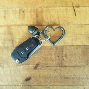 Pode incluir: Uma chave de carro preta com um chaveiro de metal em forma de coração. O chaveiro é feito de metal escuro e tem um pequeno amuleto de bolota.
