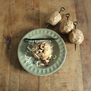 May include: A green ceramic plate with a baked potato topped with shredded cheese and bacon bits. Three potatoes are shown in the background, each with a metal skewer inserted through them.
