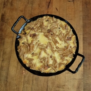 May include: A black cast iron skillet filled with a creamy pasta dish topped with melted cheese. The pasta is cooked al dente and has a golden brown colour.