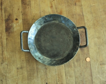 May include: A large, round, black metal pan with two handles. The pan has a slightly hammered finish and is sitting on a wooden surface.