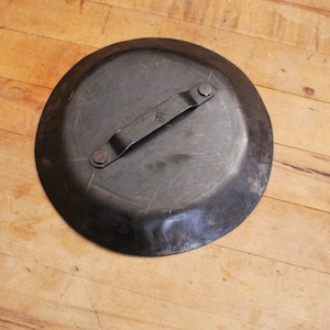 May include: A black metal plate with a handle. The plate has a slightly raised edge and a textured surface. The handle is attached to the plate with two rivets. The plate has the number "5" stamped on it.