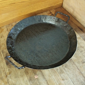 May include: A large, round, black cast iron pan with two twisted metal handles. The pan is sitting on a wooden surface.
