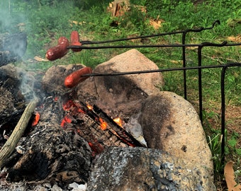 May include: Three hot dogs are roasting over a campfire on metal skewers. The campfire is surrounded by rocks and there is smoke rising from the fire.
