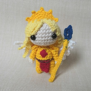 Puede incluir: Una muñeca de crochet con cabello amarillo, una corona blanca y un atuendo amarillo y rojo. La muñeca sostiene un bastón azul y amarillo.