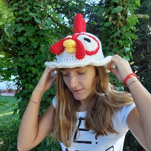 Puede incluir: Un gorro blanco de ganchillo con forma de gallo con detalles rojos y amarillos. El gorro lo lleva puesto una persona con el pelo largo y castaño.