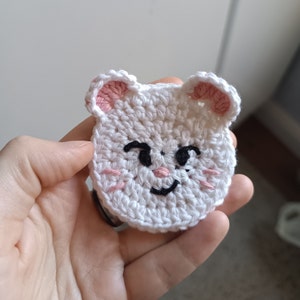 Può includere: Un volto di gatto bianco all'uncinetto con interno orecchie rosa, occhi neri e bocca sorridente. Il volto del gatto ha guance rosa ed è fatto a mano. L'articolo è di forma piccola e rotonda, probabilmente una toppa o un'applicazione.