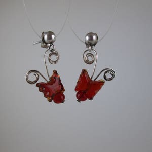 May include: A pair of dangling earrings featuring a butterfly design. The earrings have a silver-tone metal base with a red and orange butterfly-shaped pendant and a small red bead. The earrings are suspended from a silver ball stud.