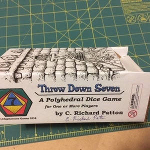 May include: A white and brown cardboard box for the game "Throw Down Seven." The box features a black and white illustration of a stone pathway and text that reads "A Polyhedral Dice Game for One or More Players." The box is on a green cutting mat.