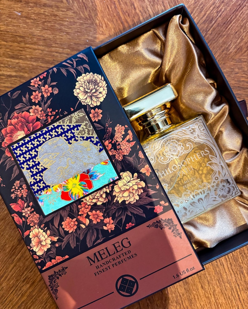 K&ouml;nnte beinhalten: Ein Parf&uuml;mflakon mit goldenem Verschluss und ge&auml;tztem Design befindet sich in einer dekorativen Schachtel. Die Schachtel ist schwarz mit floralen Mustern und der Name "MELEG" ist auf der Vorderseite aufgedruckt. Die Flasche ist 47 ml.
