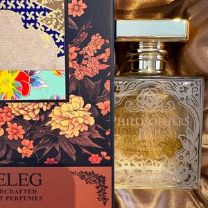 K&ouml;nnte beinhalten: Ein Parf&uuml;mflakon mit goldfarbenem Verschluss und dekorativem Etikett. Die Flasche steht neben einer Schachtel mit floralen Designs in Orange-, Gelb- und Rott&ouml;nen. Auf der Schachtel steht auch das Wort "ELEG".