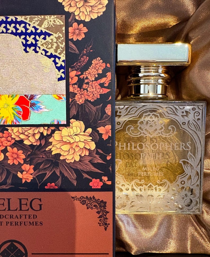 K&ouml;nnte beinhalten: Ein Parf&uuml;mflakon mit goldfarbenem Verschluss und dekorativem Etikett. Die Flasche steht neben einer Schachtel mit floralen Designs in Orange-, Gelb- und Rott&ouml;nen. Auf der Schachtel steht auch das Wort "ELEG".