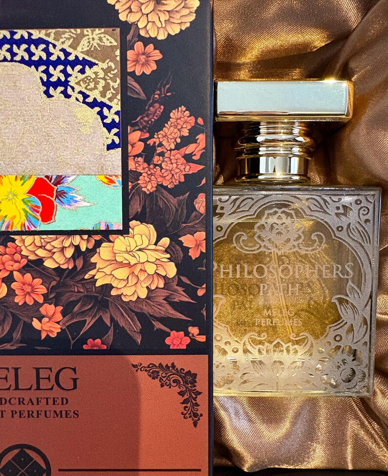 K&ouml;nnte beinhalten: Ein Parf&uuml;mflakon mit goldfarbenem Verschluss und dekorativem Etikett. Die Flasche steht neben einer Schachtel mit floralen Designs in Orange-, Gelb- und Rott&ouml;nen. Auf der Schachtel steht auch das Wort "ELEG".