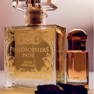 K&ouml;nnte beinhalten: Zwei Parf&uuml;mflaschen, eine gro&szlig;e und quadratische mit einem klaren, facettierten Verschluss und eine kleinere, abgerundete Flasche. Die gro&szlig;e Flasche hat goldfarbene Schrift mit der Aufschrift "PHILOSOPHERS PATH MELEG PERFUMES". Beide Flaschen sind mit einer goldenen Fl&uuml;ssigkeit gef&uuml;llt.