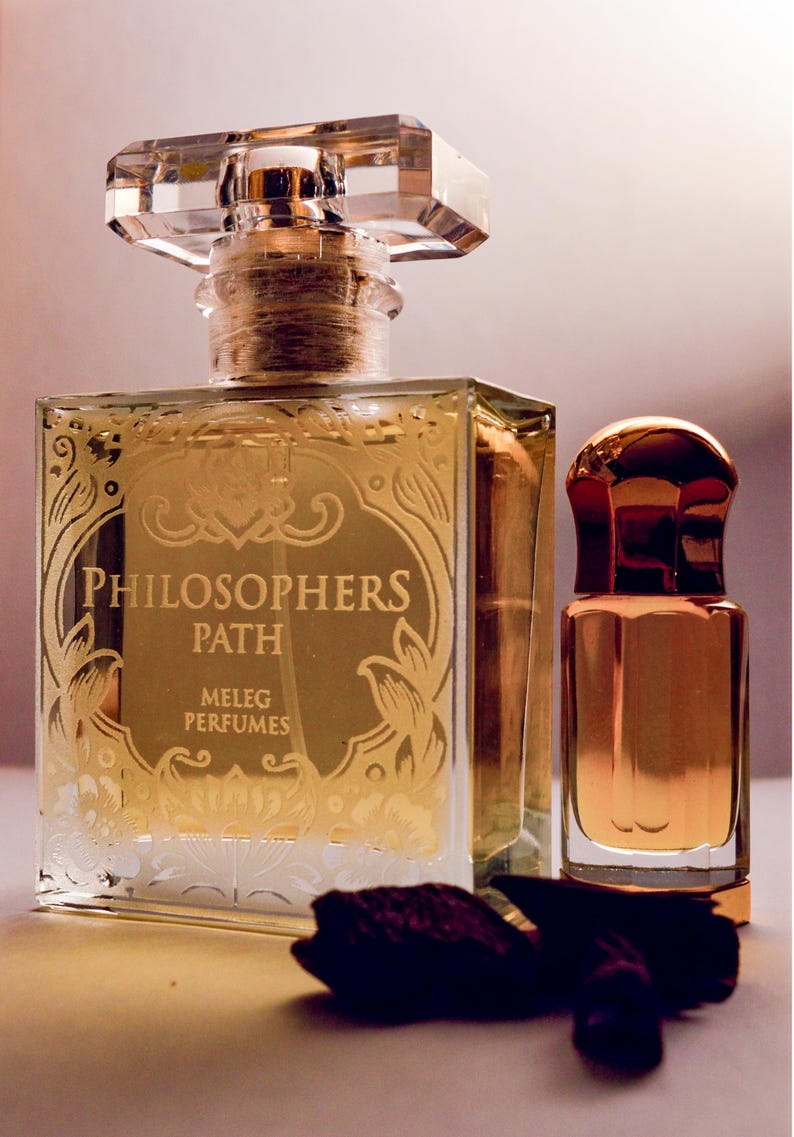 K&ouml;nnte beinhalten: Zwei Parf&uuml;mflaschen, eine gro&szlig;e und quadratische mit einem klaren, facettierten Verschluss und eine kleinere, abgerundete Flasche. Die gro&szlig;e Flasche hat goldfarbene Schrift mit der Aufschrift "PHILOSOPHERS PATH MELEG PERFUMES". Beide Flaschen sind mit einer goldenen Fl&uuml;ssigkeit gef&uuml;llt.