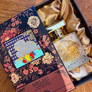 K&ouml;nnte beinhalten: Ein Parf&uuml;mflakon mit goldenem Verschluss und ge&auml;tzten Details liegt in einer offenen Schachtel. Die Schachtel ist schwarz mit floralen Designs und der Name "MELEG" ist auf der Vorderseite aufgedruckt. Die Flasche hat 47,3 ml.