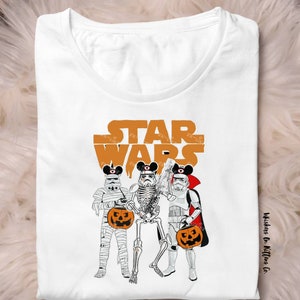 May include: White t-shirt featuring a Halloween-themed Star Wars design. The design includes three Stormtrooper figures dressed as a mummy, skeleton, and vampire, each holding a jack-o'-lantern. The words "STAR WARS" are printed in orange above the figures.