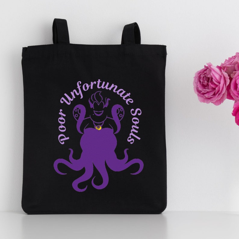 Może przedstawiać: Czarna torba typu tote z fioletowym siluetem Urszuli z bajki Mała Syrenka. Tekst "Poor Unfortunate Souls" jest napisany w k&oacute;łku wok&oacute;ł Urszuli.