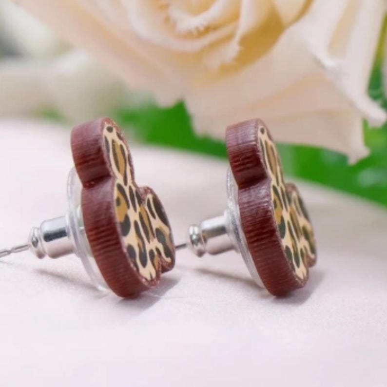 Puede incluir: Pendientes de bot&oacute;n con borde de madera marr&oacute;n y estampado de leopardo. Los pendientes tienen un poste y un cierre. El fondo incluye una flor blanca borrosa y hojas verdes. Un accesorio elegante.