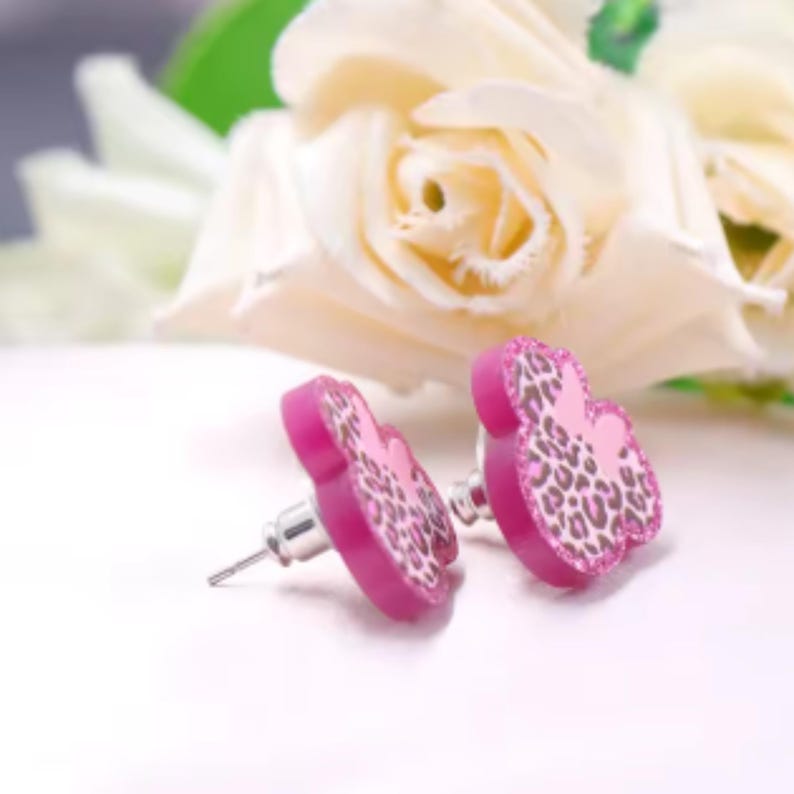 Puede incluir: Un par de pendientes de bot&oacute;n rosas con forma de cabeza de rat&oacute;n. Los pendientes tienen un dise&ntilde;o de estampado de leopardo y postes plateados. El fondo presenta rosas blancas.