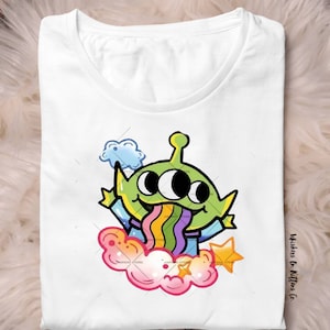 May include: White t-shirt featuring a cartoon alien with three eyes, a green head, and a rainbow-colored tongue. The alien is surrounded by a cloud, a star, and the text "Whiskers On Kittens Co."