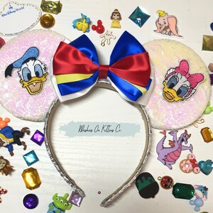 May include: A sequined headband with a large red, blue, and yellow bow. The headband has two white sequined ears with embroidered cartoon characters. Assorted Disney-themed charms and gems surround the headband.