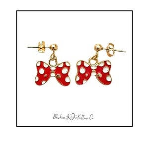 May include: A pair of gold-colored stud earrings featuring red bow charms with white polka dots. The earrings are in a bow shape and are suspended from a small gold ball. The text "Whiskers @ Kittens Co." is at the bottom.