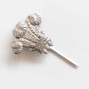 Peut inclure: Une épingle à revers en métal argenté représentant l'emblème des plumes du Prince de Galles. Le motif se compose de trois plumes d'autruche émergeant d'une base en forme de couronne, avec une longue épingle droite et pointue à la base.