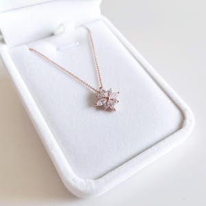 May include: A rose gold necklace with a flower-shaped pendant, set with clear crystals, is displayed in a white jewellery box. The necklace is delicate and elegant. The box is open, revealing the necklace against a soft, white interior, ideal for gifting.