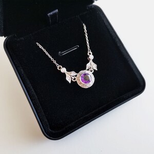 Peut inclure: Un collier en argent avec un pendentif rond en pierre précieuse violette, entouré de petites pierres blanches, présenté dans un écrin de velours noir. Le collier a une chaîne délicate et des accents en forme de feuilles.