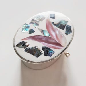 May include: Oval silver trinket box with a white enamel top. The top is decorated with iridescent abalone shell fragments and two pink, leaf-shaped pieces. A small metal loop is attached to the side.