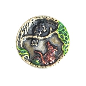 May include: A round, metal button with a detailed forest scene. A red fox is at the bottom, a black bird on a branch above, and green foliage on the right. The button has a gold-colored border.
