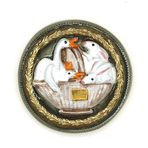 May include: A round, decorative button featuring a basket with white geese and rabbits. The button has a gold-colored border with a woven design and a gold-colored inner border. The geese and rabbits are white with orange beaks and feet.