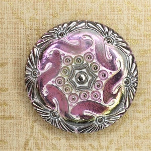 May include: A round, silver-coloured button with a pink iridescent design. The button has a raised, star-shaped centre with a smaller, silver-coloured button in the middle. The button is surrounded by a ring of smaller, silver-coloured buttons.