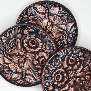 May include: Three round copper-colored ceramic coasters with an embossed floral and butterfly design. The coasters have a black background and a glossy finish.