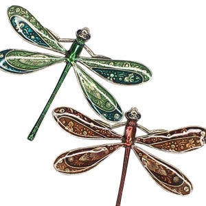 May include: Two decorative dragonfly brooches. One is green and teal, the other is brown and red. Both have detailed wing patterns and silver-colored metal accents. The brooches are set against a white background.