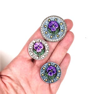 May include: Three round, decorative buttons featuring a purple pansy flower with green leaves. The buttons have silver and gold accents, with a light blue and white border. The buttons are held in a hand.
