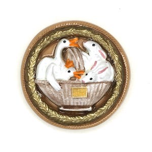 May include: A round, decorative button featuring a basket with two white geese and two white rabbits. The basket is surrounded by a gold-colored wreath and a brown border. The geese have orange beaks and feet.