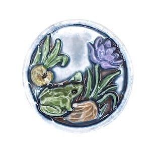 May include: A round ceramic plate with a frog, water plants, and a purple flower design. The frog is green, the flower is purple, and the plate has a blue rim. The design is raised and textured.