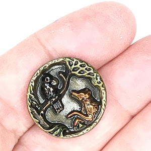 May include: A round, antique-style button with a detailed design. The button features a black bird and a copper-colored fox facing each other, framed by a tree and decorative border. The button is held in a hand.